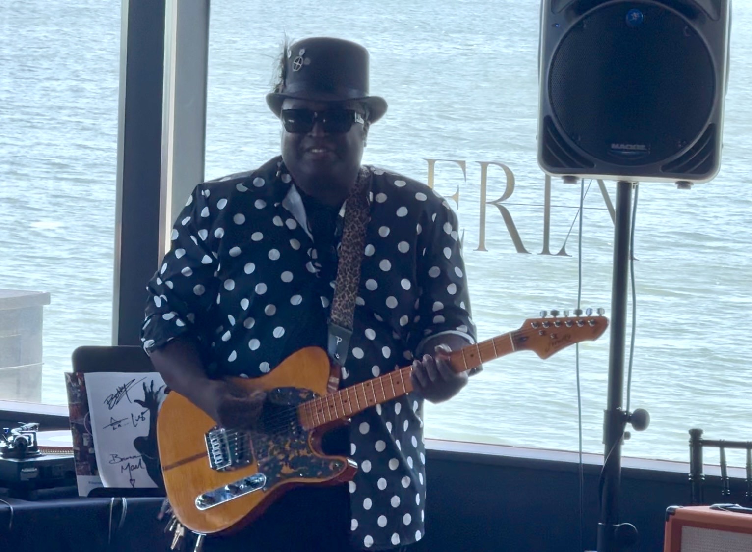 John Wesley Payne playing a natural-finish Telecaster at ERIA in a black-and-white polka-dot suit and top hat, a Prince portrait on the stand beside him and the bay through the windows