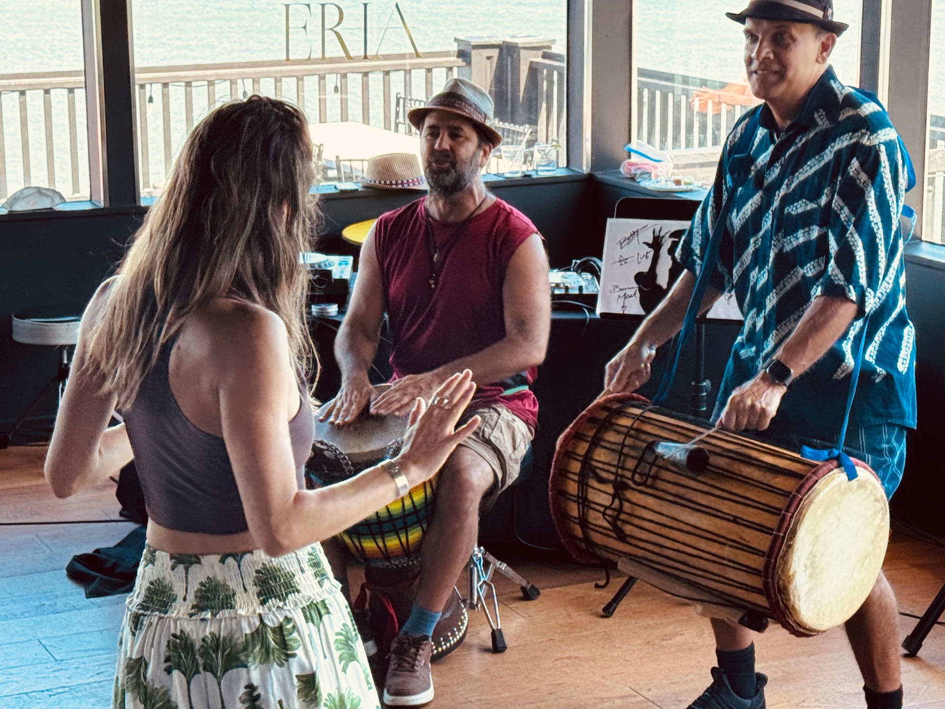 A dancer grooves as Wade Peterson and Gabriel Harris play djembe and dunun on the floor at ERIA, bay visible through the windows